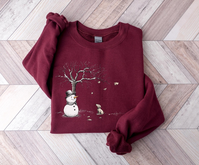 May include: A maroon sweatshirt featuring a winter scene. A snowman wearing a black top hat and scarf stands near a bare tree with snow. A small dog sits nearby, and snowflakes are falling. The sweatshirt is made of a soft material.