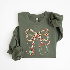 Op de afbeelding: Olijfgroene sweatshirt met een kerstthema stethoscoop ontwerp. De stethoscoop is versierd met een zuurstokstrik, zuurstokken en sneeuwvlokken. Het ontwerp is in de kleuren rood, wit en groen.