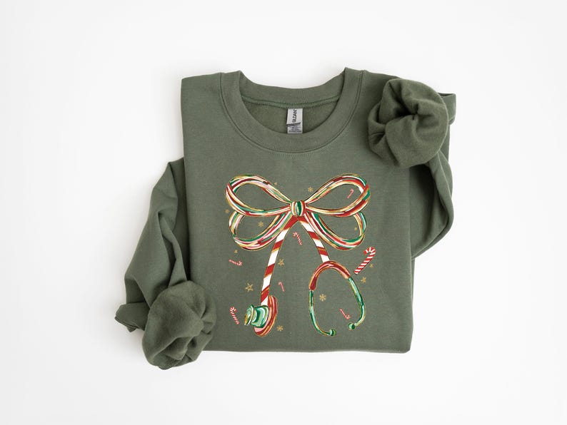 Op de afbeelding: Olijfgroene sweatshirt met een kerstthema stethoscoop ontwerp. De stethoscoop is versierd met een zuurstokstrik, zuurstokken en sneeuwvlokken. Het ontwerp is in de kleuren rood, wit en groen.