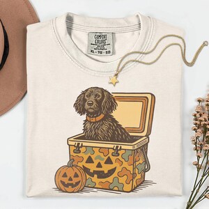 May include: Cream-colored Comfort Colors t-shirt featuring a dog inside a cooler with a jack-o'-lantern design. A gold star pendant necklace and a brown hat are also visible. The shirt has a tag that reads XL-TG-EG.