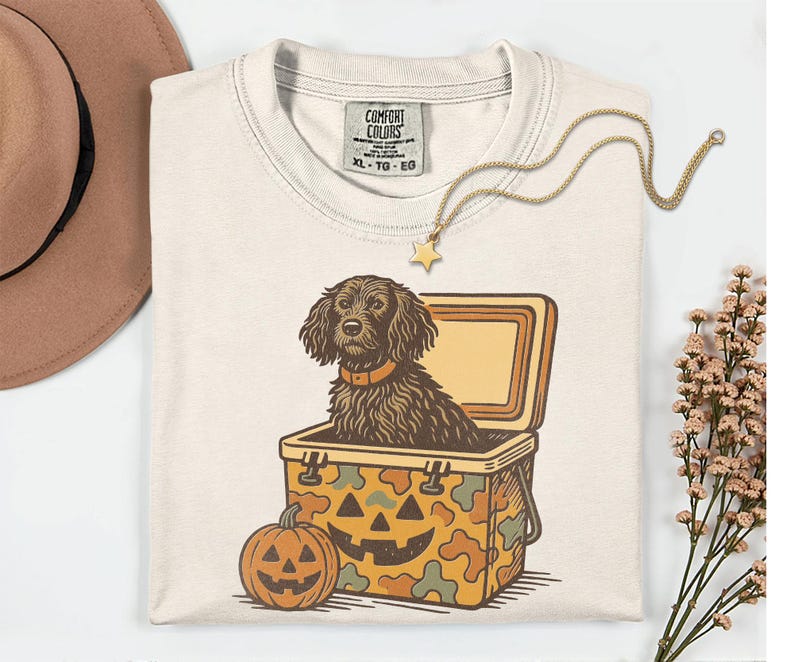 May include: Cream-colored Comfort Colors t-shirt featuring a dog inside a cooler with a jack-o'-lantern design. A gold star pendant necklace and a brown hat are also visible. The shirt has a tag that reads XL-TG-EG.