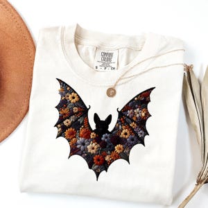 Puede incluir: Camiseta Comfort Colors color crema con un diseño de murciélago negro lleno de flores coloridas. Las alas del murciélago están adornadas con patrones florales naranjas, amarillos, morados y azules. Un collar dorado con un colgante descansa sobre la camisa.