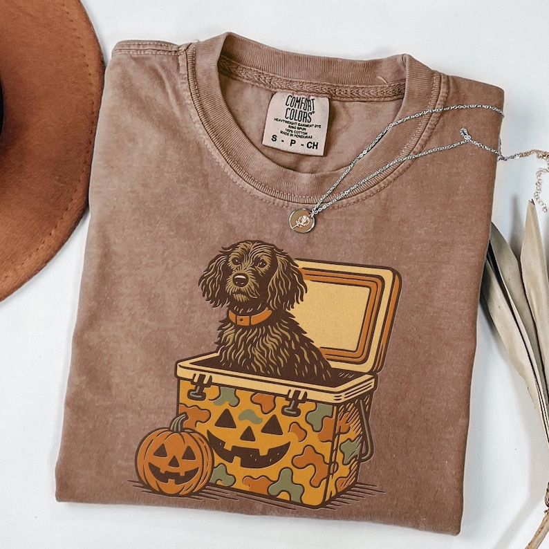 May include: Brown Comfort Colors t-shirt featuring a dog inside a cooler with a jack-o'-lantern design. The shirt has a crew neck and short sleeves. A silver necklace with a pendant is draped across the shirt.