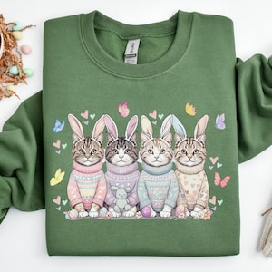 May include: Green sweatshirt featuring a graphic of four cats wearing bunny ears and sweaters. The cats are surrounded by butterflies and hearts. Easter-themed decor includes a basket of eggs and a carrot.