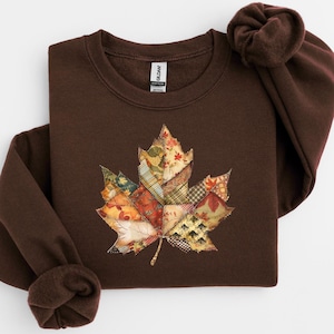 Könnte beinhalten: Ein braunes Sweatshirt mit einem großen Ahornblatt-Design im Patchwork-Stil. Das Blatt besteht aus verschiedenen Stoffmustern in Herbstfarben wie Orange, Rot und Gold. Das Sweatshirt hat einen Rundhalsausschnitt und lange Ärmel.
