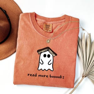 Op de afbeelding: Koraalrood Comfort Colors T-shirt met een cartoon spook met een boek op zijn hoofd. Het spook heeft zwarte ogen en een gebogen mond. De tekst "read more boooooks" staat eronder. Een gouden ketting ligt over het shirt.
