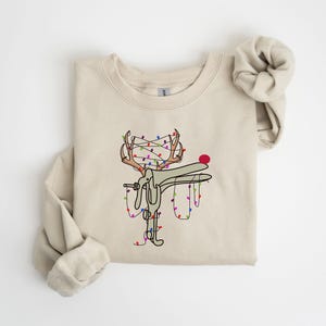 May include: A beige sweatshirt with a graphic of a reindeer with Christmas lights wrapped around its antlers. The reindeer is holding a string of lights in its mouth.