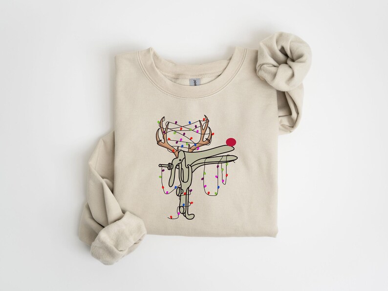 May include: A beige sweatshirt with a graphic of a reindeer with Christmas lights wrapped around its antlers. The reindeer is holding a string of lights in its mouth.
