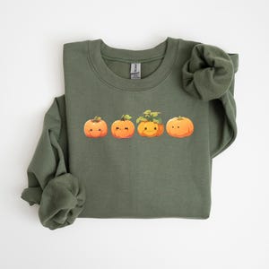 Puede incluir: Una sudadera verde oliva con una fila de cuatro calabazas de dibujos animados con caras sonrientes. Las calabazas son naranjas con tallos y hojas verdes. La sudadera tiene cuello redondo y mangas largas.