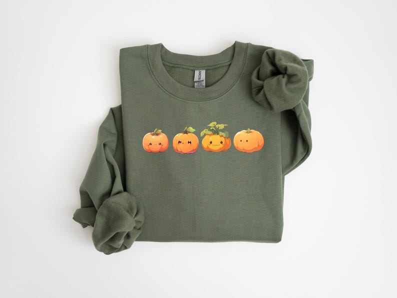 Puede incluir: Una sudadera verde oliva con una fila de cuatro calabazas de dibujos animados con caras sonrientes. Las calabazas son naranjas con tallos y hojas verdes. La sudadera tiene cuello redondo y mangas largas.