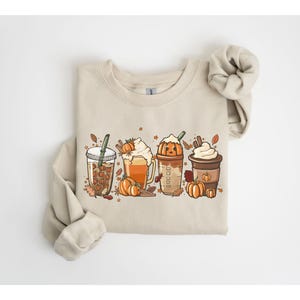 May include: A beige sweatshirt with a graphic of four autumn-themed drinks: iced coffee, pumpkin spice latte, pumpkin spice frappe, and a whipped cream coffee. The design includes pumpkins, leaves, and cinnamon sticks. The sweatshirt has long sleeves and a crew neck.