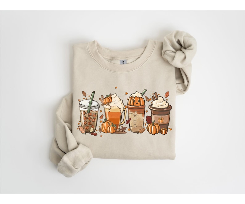 May include: A beige sweatshirt with a graphic of four autumn-themed drinks: iced coffee, pumpkin spice latte, pumpkin spice frappe, and a whipped cream coffee. The design includes pumpkins, leaves, and cinnamon sticks. The sweatshirt has long sleeves and a crew neck.