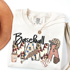 May include: Cream-colored t-shirt with the words "Baseball MAMA" in a baseball-themed design. The design includes a baseball bat, baseballs, a baseball glove, and a baseball helmet. A gold necklace and a brown hat are also visible.