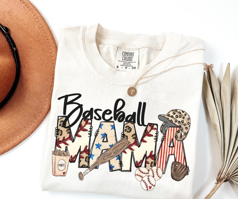 May include: Cream-colored t-shirt with the words "Baseball MAMA" in a baseball-themed design. The design includes a baseball bat, baseballs, a baseball glove, and a baseball helmet. A gold necklace and a brown hat are also visible.