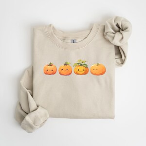Puede incluir: Sudadera beige con un dise&ntilde;o de cuatro calabazas de dibujos animados en fila. Las calabazas naranjas tienen caras sonrientes y tallos verdes. La sudadera tiene mangas largas y cuello redondo.