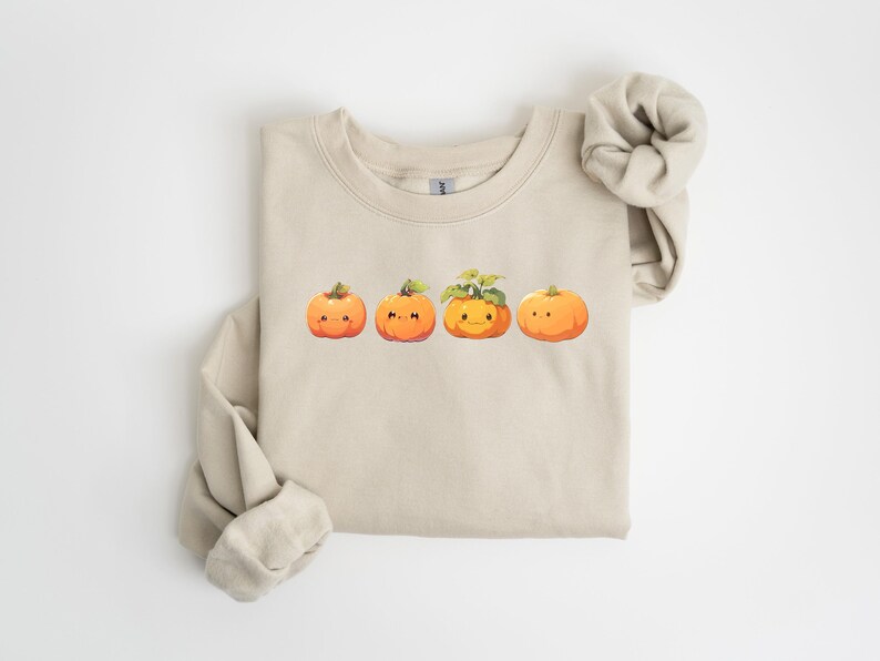 Puede incluir: Sudadera beige con un dise&ntilde;o de cuatro calabazas de dibujos animados en fila. Las calabazas naranjas tienen caras sonrientes y tallos verdes. La sudadera tiene mangas largas y cuello redondo.