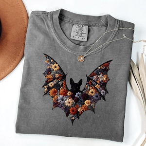 K&ouml;nnte beinhalten: Ein graues Comfort Colors T-Shirt mit einem floralen Fledermaus-Design. Die Fl&uuml;gel der Fledermaus sind mit bunten Blumen in Orange-, Gelb- und Lilat&ouml;nen gef&uuml;llt. Eine silberne Halskette mit einem kleinen Anh&auml;nger liegt auf dem T-Shirt.
