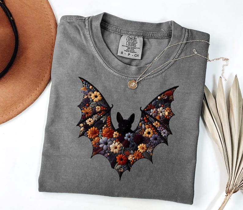 K&ouml;nnte beinhalten: Ein graues Comfort Colors T-Shirt mit einem floralen Fledermaus-Design. Die Fl&uuml;gel der Fledermaus sind mit bunten Blumen in Orange-, Gelb- und Lilat&ouml;nen gef&uuml;llt. Eine silberne Halskette mit einem kleinen Anh&auml;nger liegt auf dem T-Shirt.
