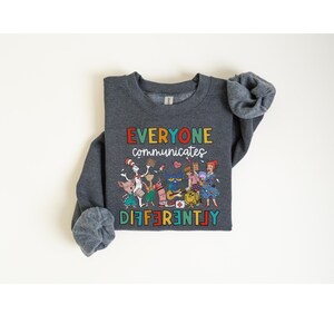 May include: A dark gray sweatshirt with a colorful graphic of characters from children's books. The text "EVERYONE communicates DIFFERENTLY" is printed in rainbow colors on the sweatshirt.