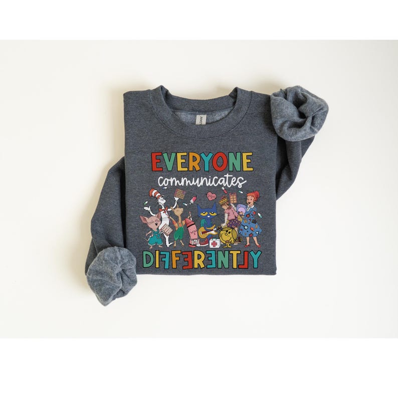 May include: A dark gray sweatshirt with a colorful graphic of characters from children's books. The text "EVERYONE communicates DIFFERENTLY" is printed in rainbow colors on the sweatshirt.