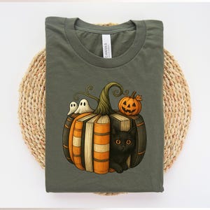 May include: Olive green t-shirt featuring a Halloween-themed graphic. The design includes a pumpkin made of books, with a black cat, two ghosts, and a jack-o'-lantern. The shirt is made of a soft material.