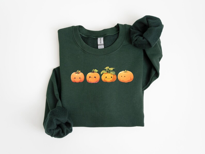 Puede incluir: Sudadera verde oscuro con cuatro calabazas de dibujos animados. Las calabazas son naranjas con hojas y tallos verdes. La sudadera tiene cuello redondo y mangas largas, ideal para el oto&ntilde;o.