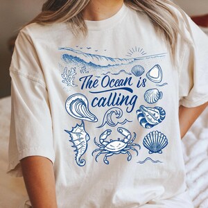 Puede incluir: Camiseta color crema con un gráfico azul con temática oceánica. El diseño incluye una ola, conchas marinas, un caballito de mar y un cangrejo, con el texto "The Ocean is calling". Una camiseta casual y cómoda para los amantes de la playa.