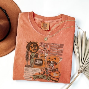 May include: A coral-colored t-shirt with a Halloween-themed graphic. The design includes a jack-o'-lantern, an alien, and the text "Trick or Treat." A gold necklace and a brown hat are also visible.