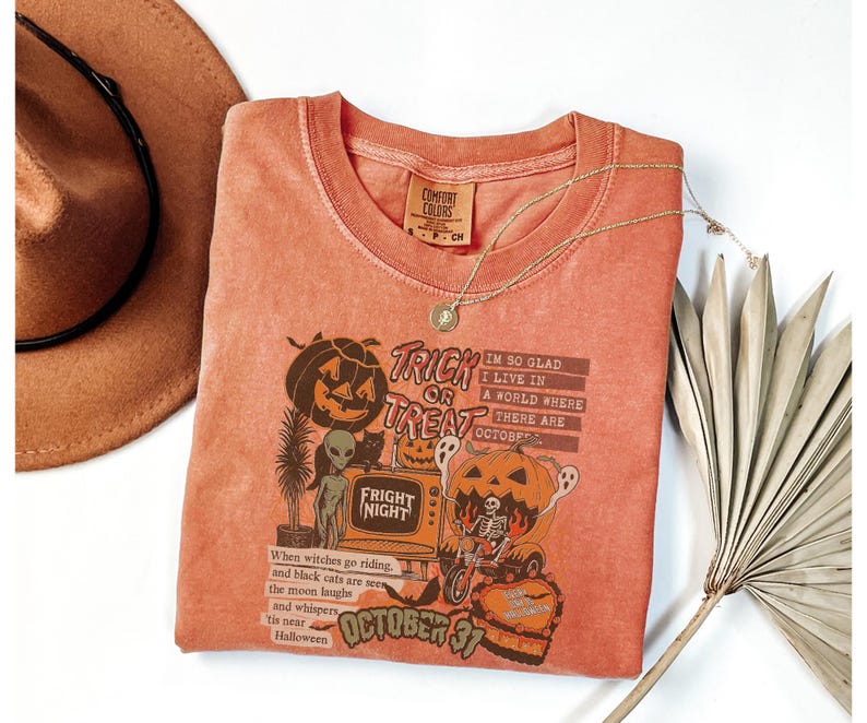 May include: A coral-colored t-shirt with a Halloween-themed graphic. The design includes a jack-o'-lantern, an alien, and the text "Trick or Treat." A gold necklace and a brown hat are also visible.