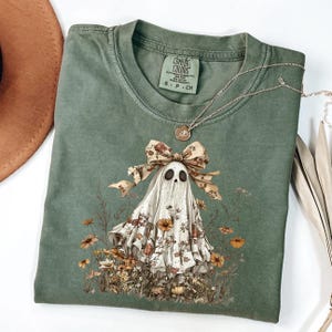 Puede incluir: Una camiseta Comfort Colors verde salvia con un diseño de fantasma floral. El fantasma tiene un lazo floral y está rodeado de flores. Un collar plateado con un colgante está drapeado sobre la camisa. Un sombrero marrón es visible en la esquina superior izquierda.
