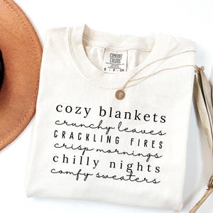 May include: Cream-colored t-shirt with the words "cozy blankets, crunchy leaves, crackling fires, crisp mornings, chilly nights, comfy sweaters" in black script. A gold necklace and a brown hat are also visible.