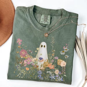 Puede incluir: Camiseta Comfort Colors verde oliva con un diseño de fantasma en un jardín floral. El fantasma es blanco con ojos negros, rodeado de flores rosas, naranjas y blancas. Un collar dorado con un colgante descansa sobre la camiseta. Talla S-P-CH.
