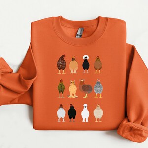 May include: Orange sweatshirt featuring a colorful illustration of various chicken breeds. The design includes a variety of chickens in different colors and patterns. The sweatshirt has a crew neck and long sleeves, perfect for casual wear.