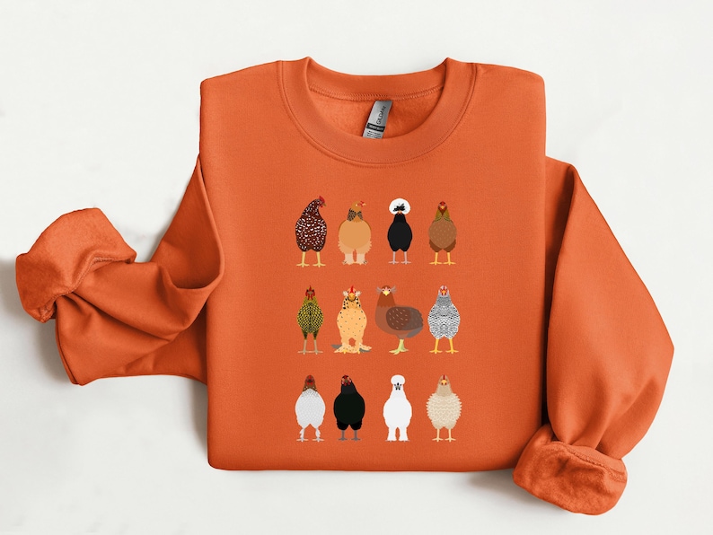 May include: Orange sweatshirt featuring a colorful illustration of various chicken breeds. The design includes a variety of chickens in different colors and patterns. The sweatshirt has a crew neck and long sleeves, perfect for casual wear.
