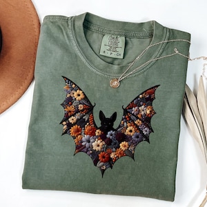 K&ouml;nnte beinhalten: Salbeigr&uuml;nes T-Shirt mit floralem Fledermaus-Design. Die Fledermausfl&uuml;gel sind mit bunten Blumen in Orange-, Gelb- und Lilat&ouml;nen gef&uuml;llt. Eine silberne Halskette mit einem kleinen Anh&auml;nger liegt auf dem T-Shirt. Das T-Shirt hat ein Comfort Colors-Etikett.