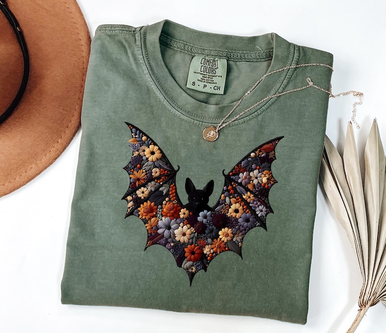 K&ouml;nnte beinhalten: Salbeigr&uuml;nes T-Shirt mit floralem Fledermaus-Design. Die Fledermausfl&uuml;gel sind mit bunten Blumen in Orange-, Gelb- und Lilat&ouml;nen gef&uuml;llt. Eine silberne Halskette mit einem kleinen Anh&auml;nger liegt auf dem T-Shirt. Das T-Shirt hat ein Comfort Colors-Etikett.