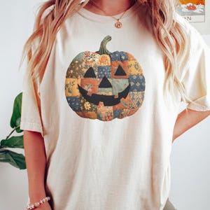 Może przedstawiać: Kremowy t-shirt z patchworkowym wzorem dyni. Dynia ma uśmiechniętą twarz i jest wykonana z r&oacute;żnych wzor&oacute;w tkanin w odcieniach pomarańczu, niebieskiego i ż&oacute;łtego. Zielona łodyga wieńczy dynię, idealną na jesień.