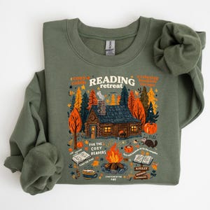 Puede incluir: Sudadera verde oliva con un gráfico de "Reading Retreat". El diseño presenta una escena de cabaña acogedora con una fogata, calabazas y libros. El texto incluye frases como "cozy cabin", "relaxing bookish escape" y "for the cozy readers".