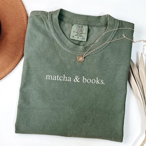 May include: An olive green t-shirt with the words "matcha & books" printed in white. A gold necklace with a pendant rests on the shirt. The shirt is folded and placed on a white surface, with a brown hat and dried leaves in the background.