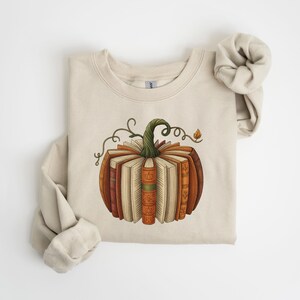 May include: Beige sweatshirt featuring a pumpkin design made of books. The pumpkin has a green stem and vine details. The books are various shades of brown and orange, with open pages. The sweatshirt's sleeves are rolled up.