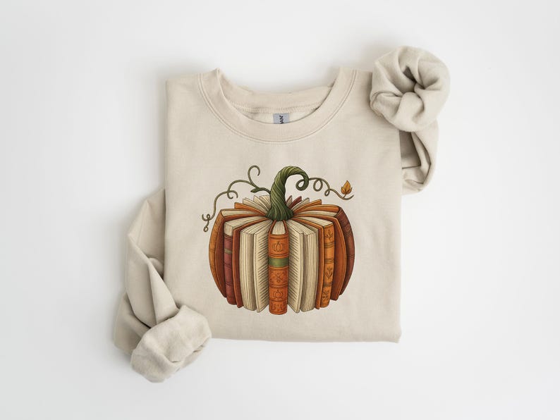 May include: Beige sweatshirt featuring a pumpkin design made of books. The pumpkin has a green stem and vine details. The books are various shades of brown and orange, with open pages. The sweatshirt's sleeves are rolled up.
