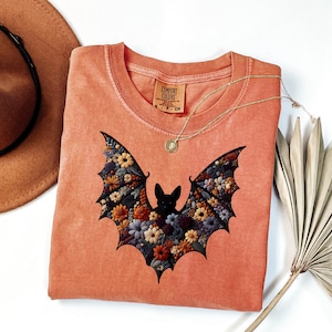K&ouml;nnte beinhalten: Korallfarbenes Comfort Colors T-Shirt mit floralem Fledermaus-Design. Die Fl&uuml;gel der Fledermaus sind mit bunten Blumen gef&uuml;llt. Eine goldene Halskette mit Anh&auml;nger liegt &uuml;ber dem Shirt. Ein brauner Hut und getrocknete Palmbl&auml;tter sind im Hintergrund.