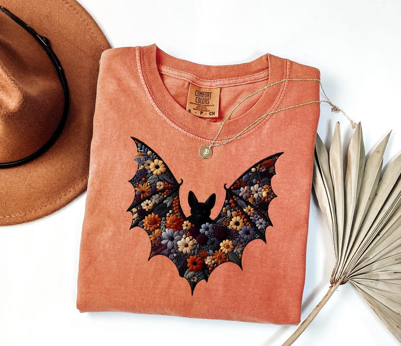 K&ouml;nnte beinhalten: Korallfarbenes Comfort Colors T-Shirt mit floralem Fledermaus-Design. Die Fl&uuml;gel der Fledermaus sind mit bunten Blumen gef&uuml;llt. Eine goldene Halskette mit Anh&auml;nger liegt &uuml;ber dem Shirt. Ein brauner Hut und getrocknete Palmbl&auml;tter sind im Hintergrund.