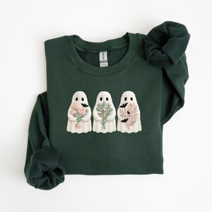 K&ouml;nnte beinhalten: Dunkelgr&uuml;nes Sweatshirt mit drei wei&szlig;en Geistermotiven, die jeweils Blumen- und Fledermausmotive halten. Die Geister haben schwarze Punktaugen. Ein lang&auml;rmliges Oberteil, perfekt f&uuml;r Halloween oder die gruselige Jahreszeit. Bequemes, l&auml;ssiges Kleidungsst&uuml;ck.