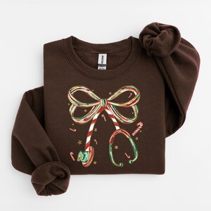 Op de afbeelding: Een donkerbruin sweatshirt met een feestelijk ontwerp. De afbeelding bevat een strik en een stethoscoop, beide versierd met zuurstokstrepen en accenten. Het ontwerp is in rood, groen en goud, met kleine sterdetails.