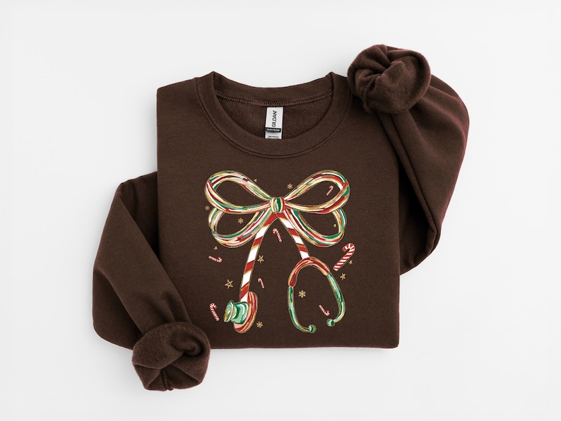 Op de afbeelding: Een donkerbruin sweatshirt met een feestelijk ontwerp. De afbeelding bevat een strik en een stethoscoop, beide versierd met zuurstokstrepen en accenten. Het ontwerp is in rood, groen en goud, met kleine sterdetails.