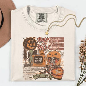 May include: Cream-colored Halloween-themed t-shirt with a graphic featuring pumpkins, ghosts, and the text "Trick or Treat" and "Fright Night." The shirt also has the text "October 31" and a quote about Halloween. A gold star pendant necklace rests on the shirt.
