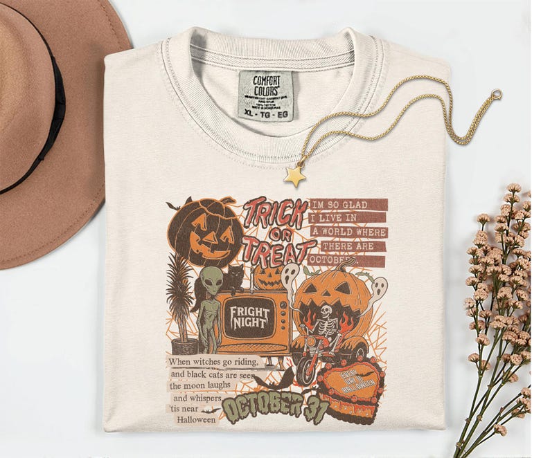 May include: Cream-colored Halloween-themed t-shirt with a graphic featuring pumpkins, ghosts, and the text "Trick or Treat" and "Fright Night." The shirt also has the text "October 31" and a quote about Halloween. A gold star pendant necklace rests on the shirt.