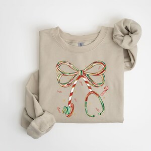Op de afbeelding: Beige sweatshirt met een kerstthema. De afbeelding toont een strik gemaakt van zuurstokken en een stethoscoop, beide versierd met zuurstokstrepen. Kleine zuurstokken zijn verspreid rond het ontwerp.
