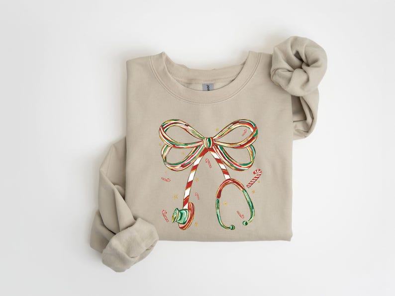 Op de afbeelding: Beige sweatshirt met een kerstthema. De afbeelding toont een strik gemaakt van zuurstokken en een stethoscoop, beide versierd met zuurstokstrepen. Kleine zuurstokken zijn verspreid rond het ontwerp.